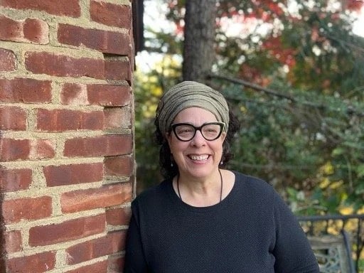 May contain a person wearing a light-gray head wrap, glasses, and a black sweater, leaning against a brick wall.