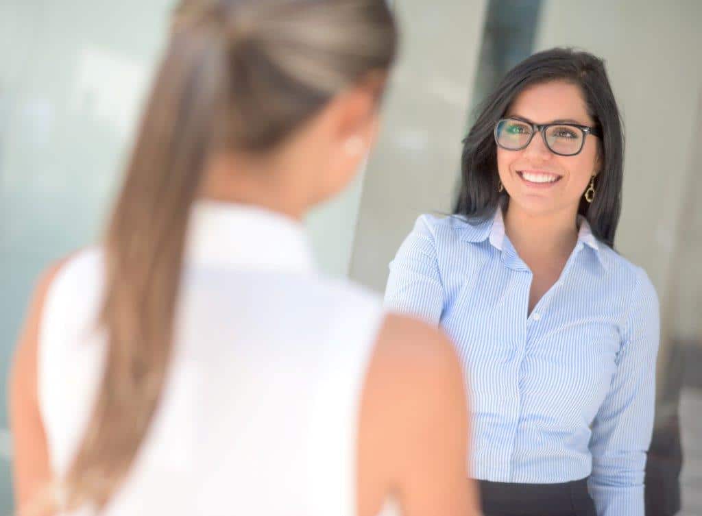 A woman with shoulder-length dark brown hair and glasses is smiling at the camera. She is wearing a black top, and greenery is visible in the blurred background.