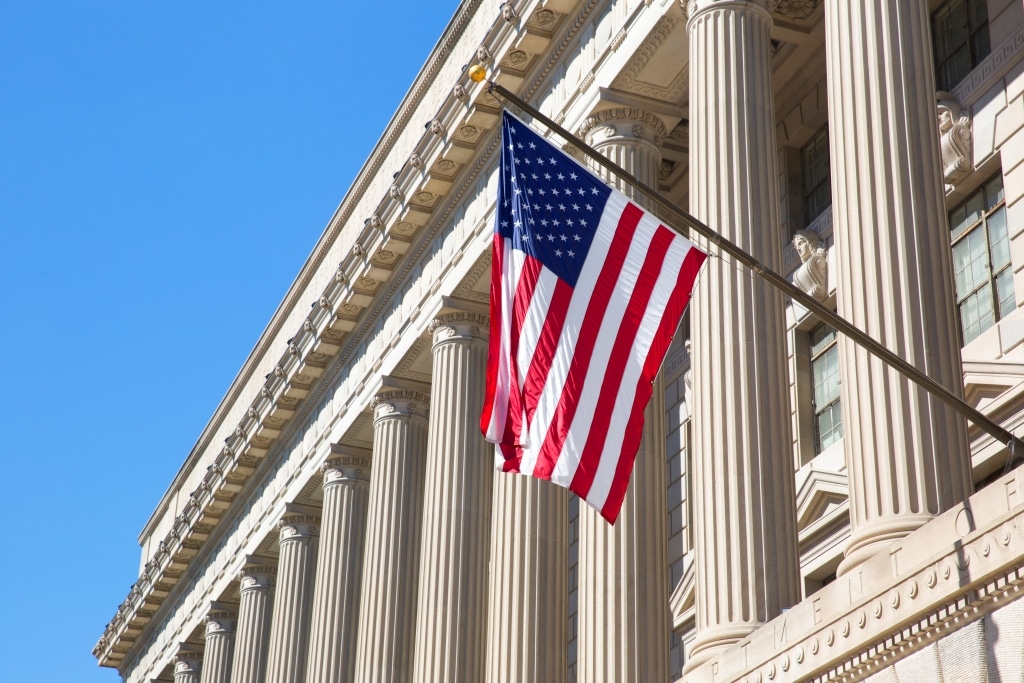 An American flag hangs on a pole in front of a large, classical-style government building with tall columns under a clear blue sky.