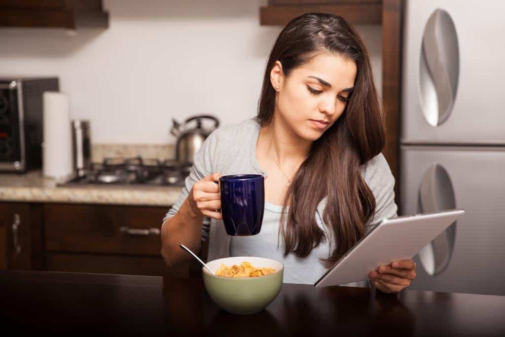A woman sits at a kitchen table holding a blue mug and looking at a tablet. A bowl of cereal with a spoon is in front of her. The kitchen has dark cabinets and a stainless steel refrigerator.
