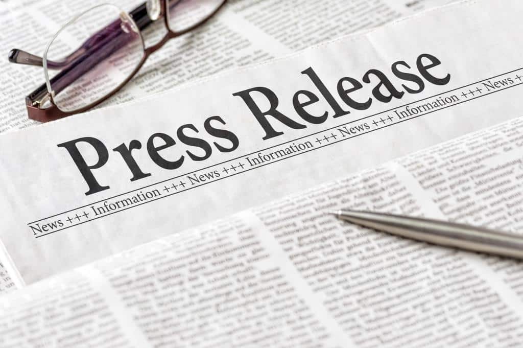 A close-up of a newspaper showing the words Press Release at the top, with eyeglasses and a pen placed on the pages. The background consists of printed news text.