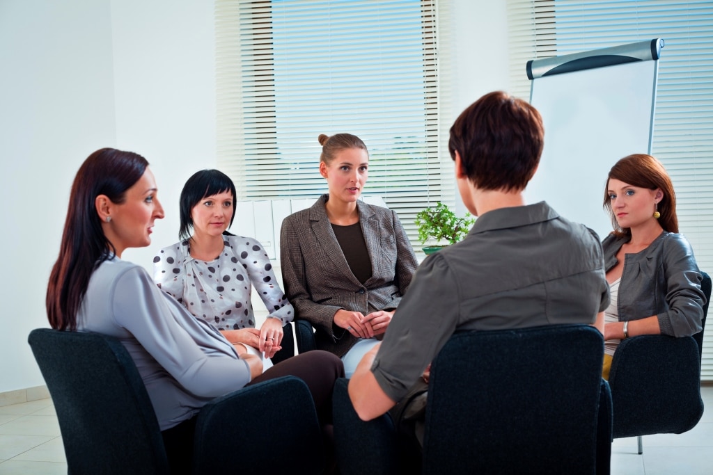 Five women sit in a circle of chairs in an office setting, engaged in a group discussion. A white flipchart and a plant are visible in the background near a window with blinds.
