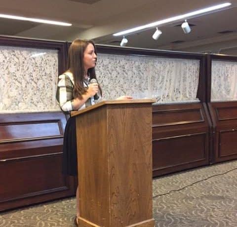 A woman stands behind a wooden podium, holding a microphone and speaking. She is indoors, with closed caskets lined up in the background. The room is well-lit with overhead lights.