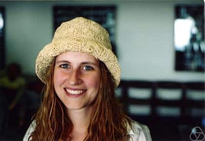 A woman with long, wavy hair wearing a beige knit hat and a white shirt smiles at the camera. The background is out of focus, showing chairs and figures indoors.