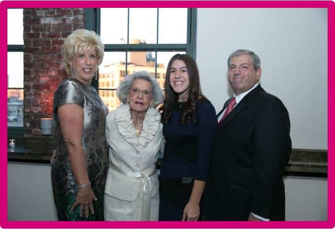 Four people stand indoors in semi-formal attire, posing for a photo in front of a window with a cityscape outside. The group includes three women and one man, all smiling. A bright pink border frames the image.