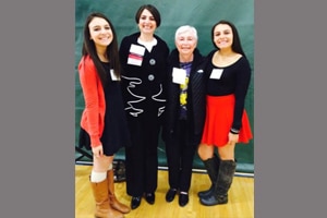 Four women stand indoors on a gym floor in front of a green backdrop, posing for a group photo. Two are wearing skirts and boots, one is in a black suit, and one has short gray hair and is wearing a dark jacket.