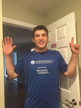 A young man stands in a doorway with both hands raised, wearing a blue T-shirt with Sharsheret and Bernstein Liebhard printed on it. The door behind him is partially open and the room is well-lit.