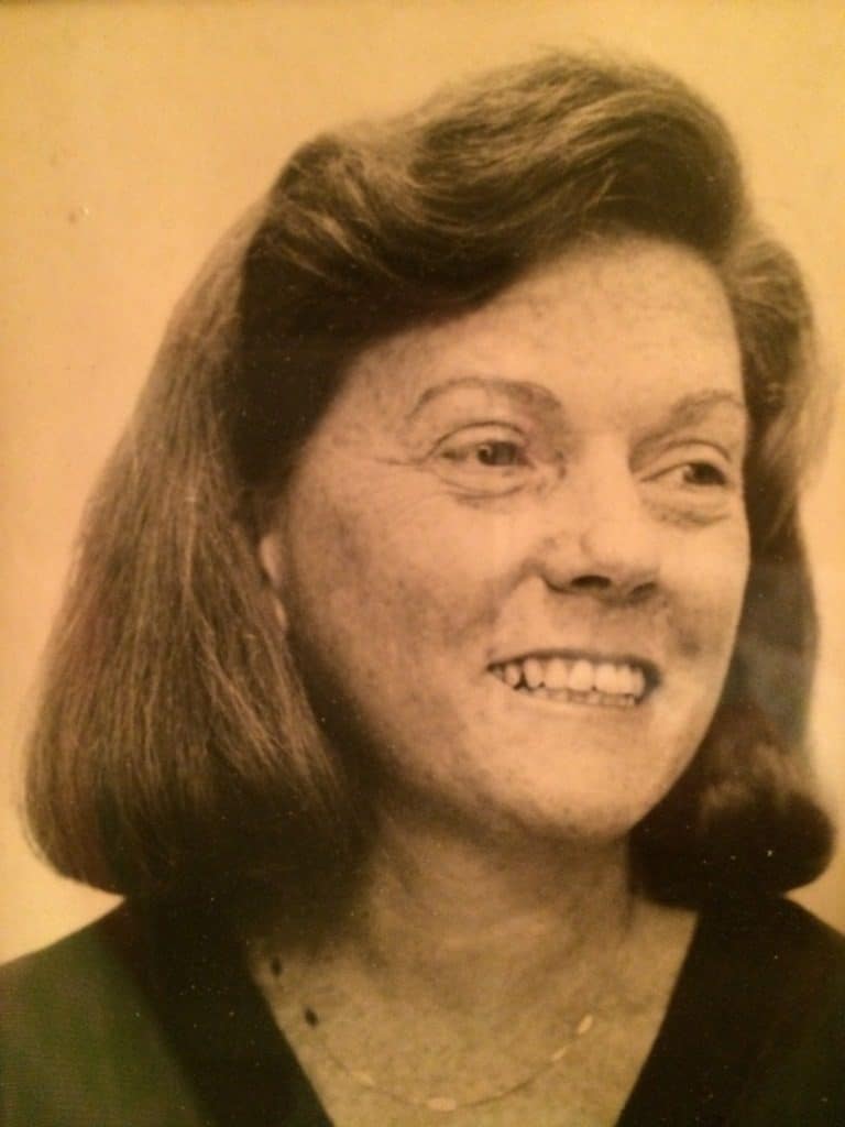 A sepia-toned portrait of a woman with shoulder-length hair, parted to the side. She is wearing a dark top and a thin necklace, and is smiling while looking slightly to her left.