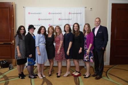 A group of eight women and one man stand in a line, smiling at the camera, in front of a white backdrop with repeating SHARSHERET logos at an indoor event.