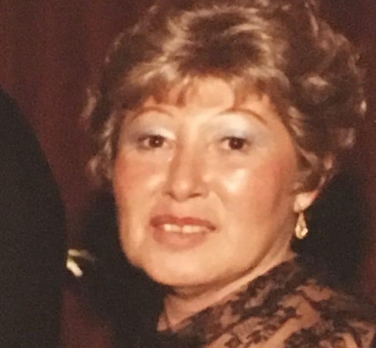 A woman with short, curly light brown hair and light makeup is wearing a pair of small gold earrings and a lace top. She is looking toward the camera with a neutral expression. The background is dark and out of focus.