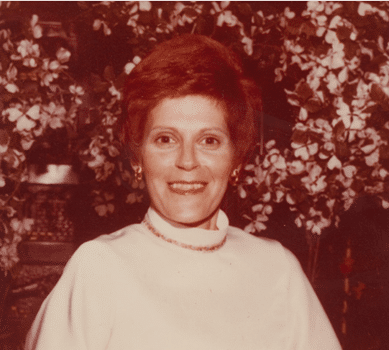 A woman with short, reddish-brown hair and a white top smiles at the camera. She is sitting in front of a background filled with flowers and greenery. The image has a warm, vintage tone.