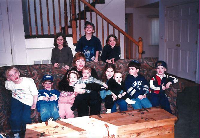 A group of twelve children pose for a photo indoors, with some sitting on a patterned couch and others on the stairs behind them. The room includes a wooden coffee table and a staircase with a wooden railing.
