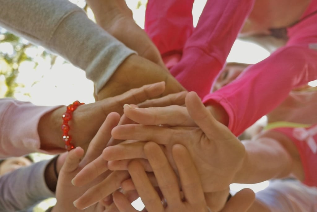A group of people stack their hands together in the center, showing teamwork or unity. The people are wearing long-sleeved shirts in light colors and pink. One person has a red bead bracelet.