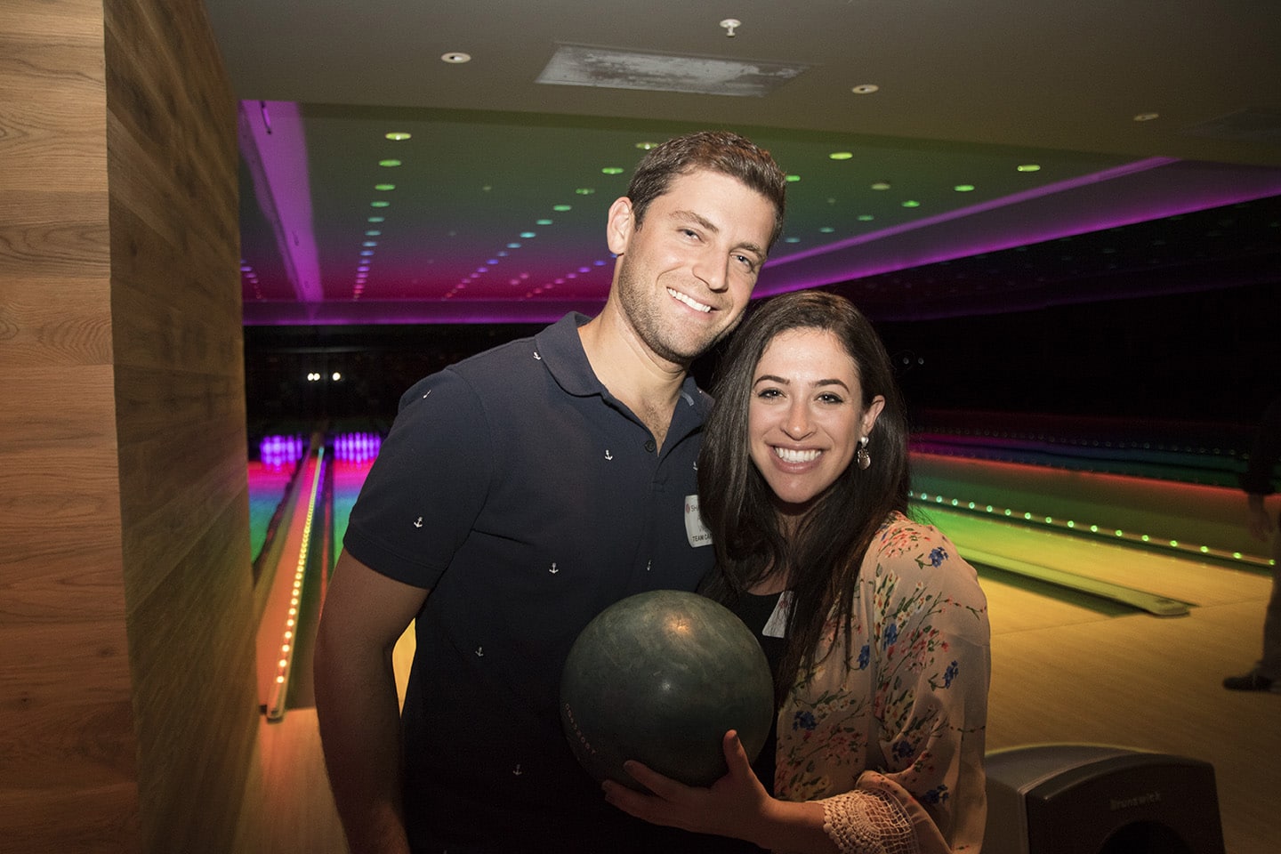 A man and a woman stand together in front of bowling lanes with colorful lights. The woman holds a bowling ball and they are both smiling at the camera.