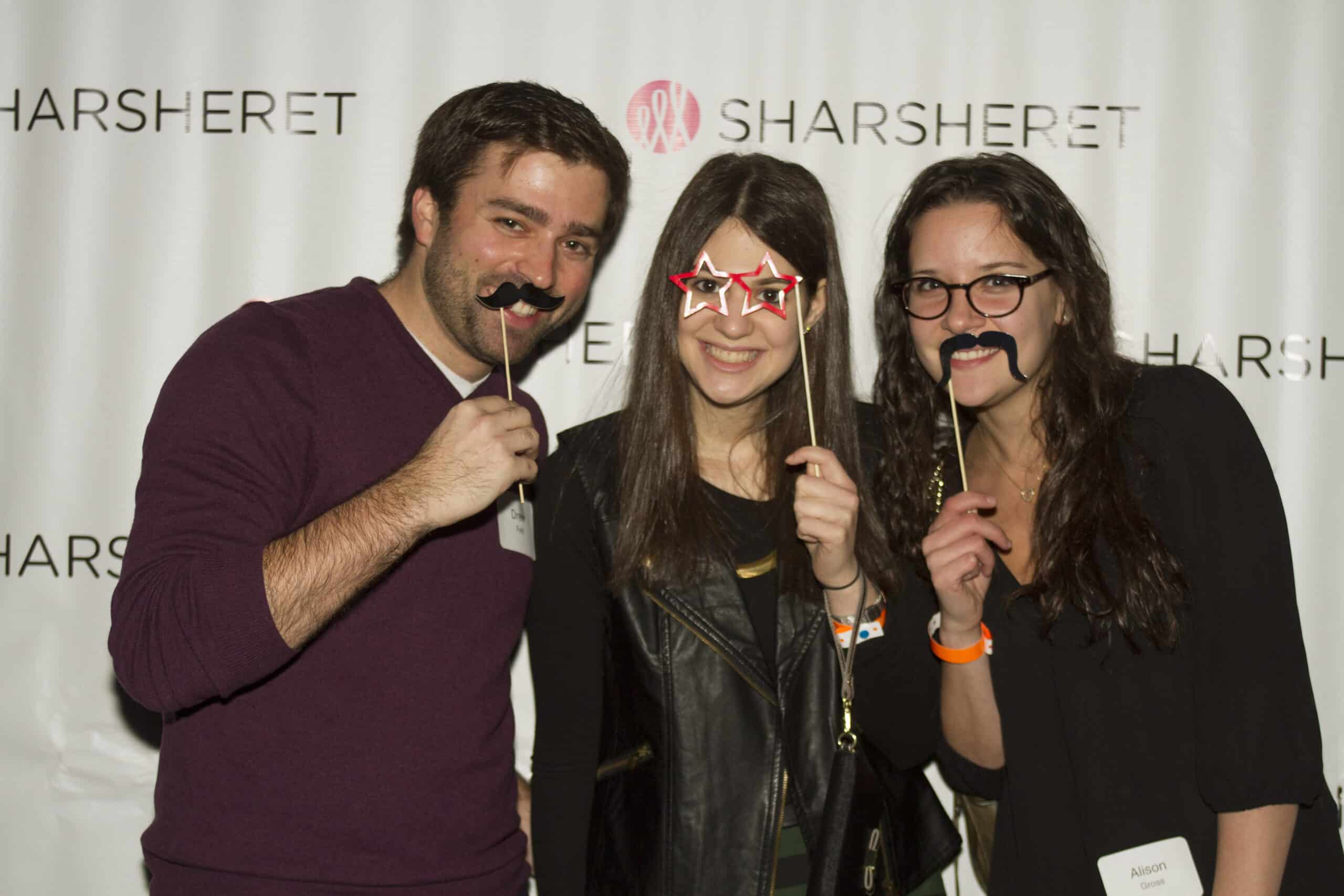 Three people stand in front of a Sharsheret backdrop, smiling and holding fun props, including star-shaped glasses and fake mustaches on sticks. Two wear name tags.