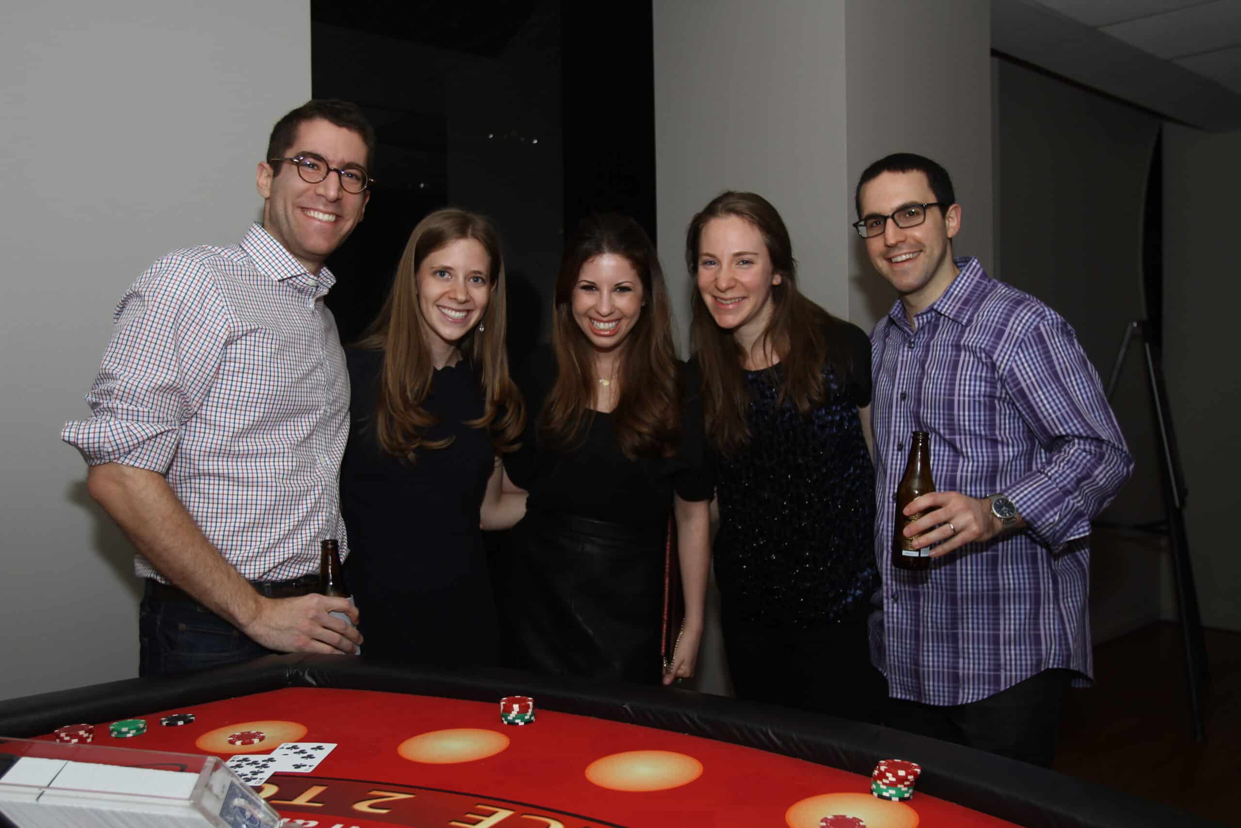 Five people stand and smile behind a casino table with playing cards, poker chips, and drinks in hand. They are indoors and casually dressed, appearing to enjoy a social gathering.