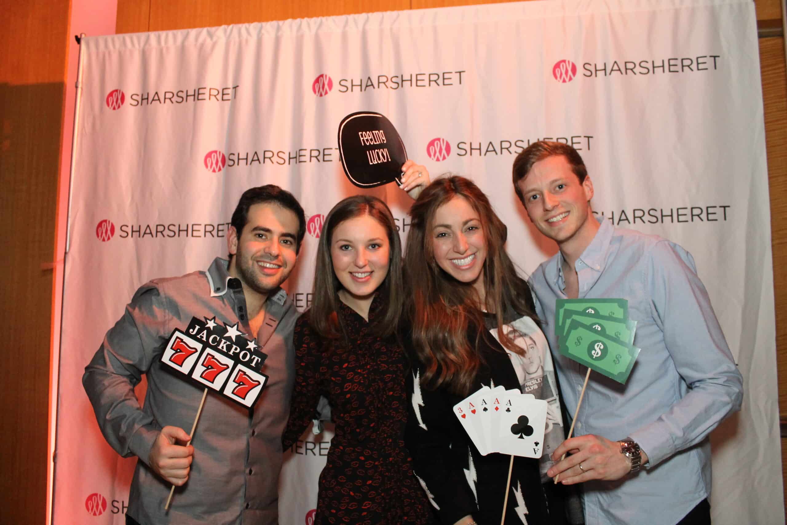 Four people stand in front of a Sharsheret step-and-repeat banner, smiling and holding casino-themed props, including play money, playing cards, a jackpot sign, and a photo booth sign.