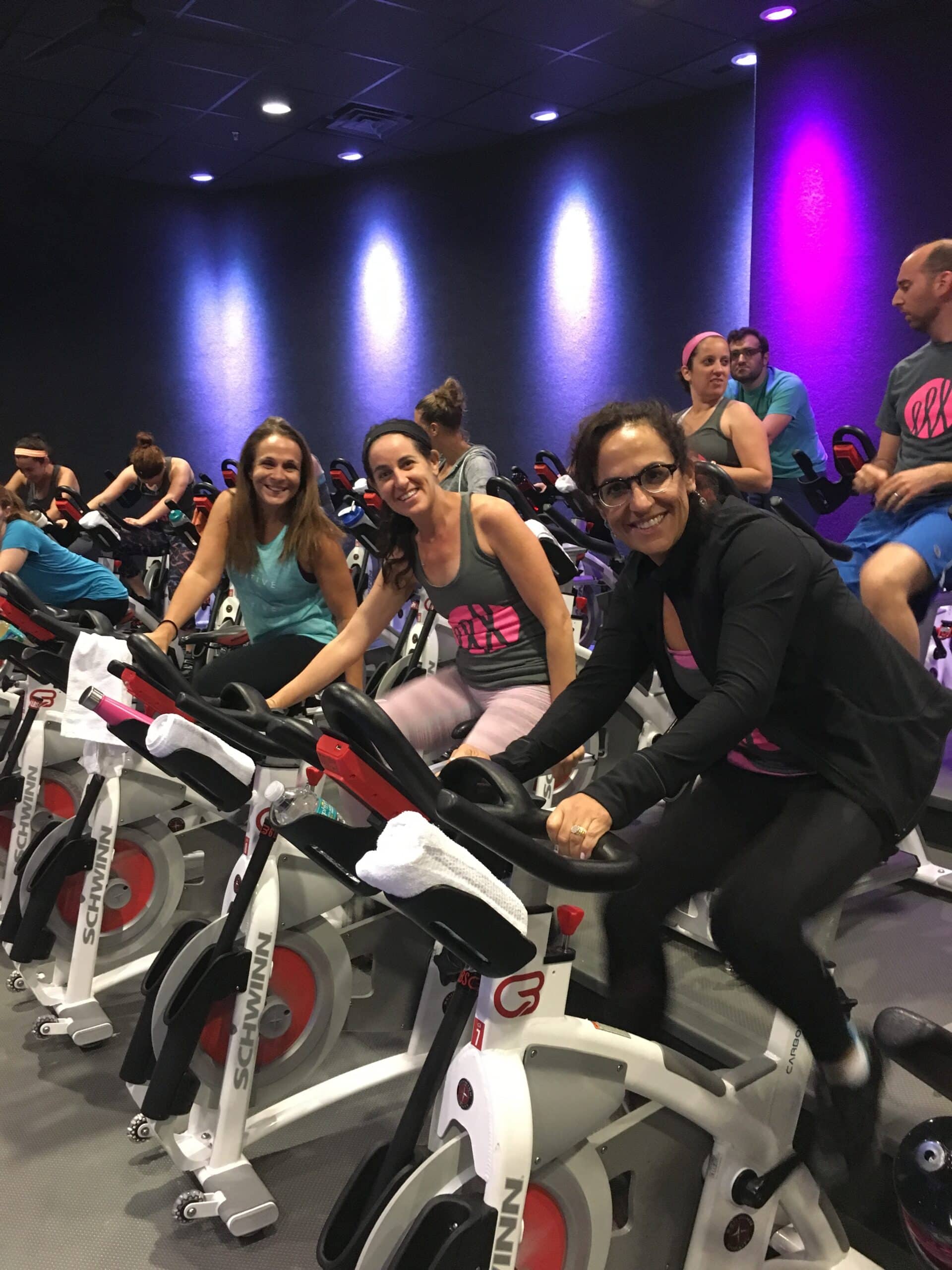 A group of people participate in an indoor cycling class. Three women in the foreground are on stationary bikes, smiling at the camera. Other participants are also cycling in the background under purple lighting.