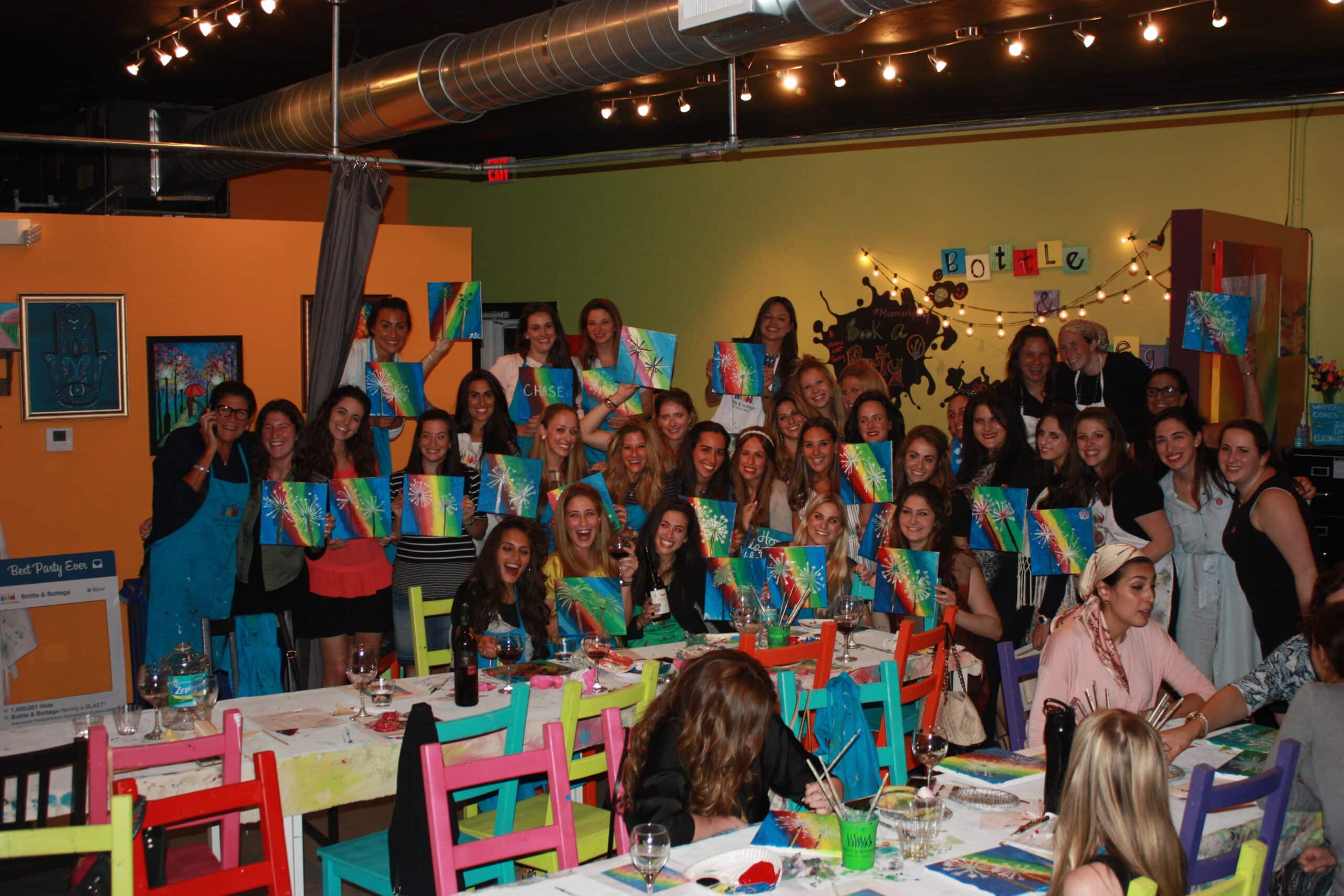 A large group of people pose together in a brightly lit, colorful art studio, each holding up their completed canvas paintings. Tables with art supplies are in front of them, and string lights hang from the ceiling.