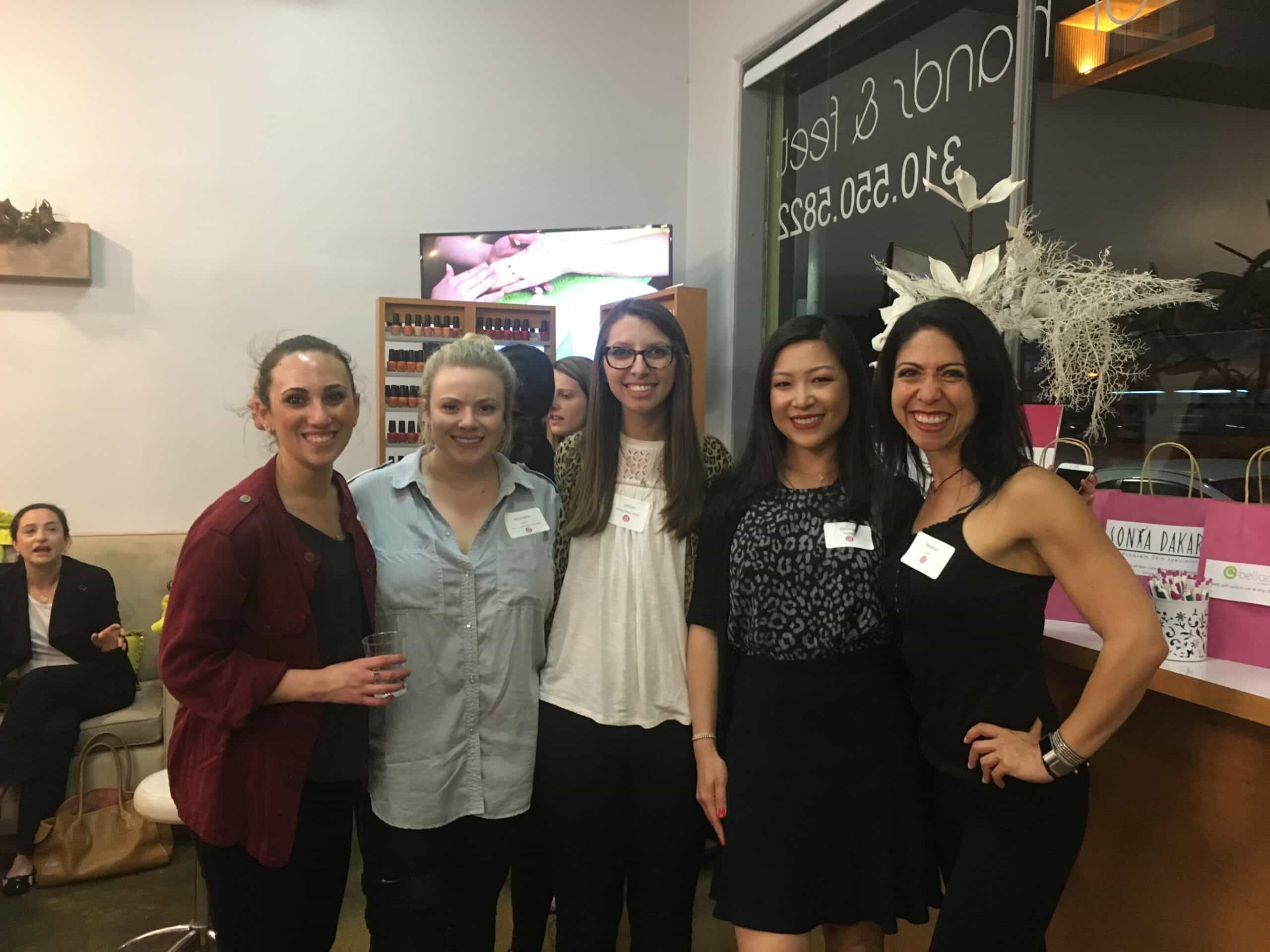 Five women stand together indoors, smiling at the camera. Four are grouped closely in the foreground, while one is in the background to the left. Gift bags and a counter are visible on the right side of the image.