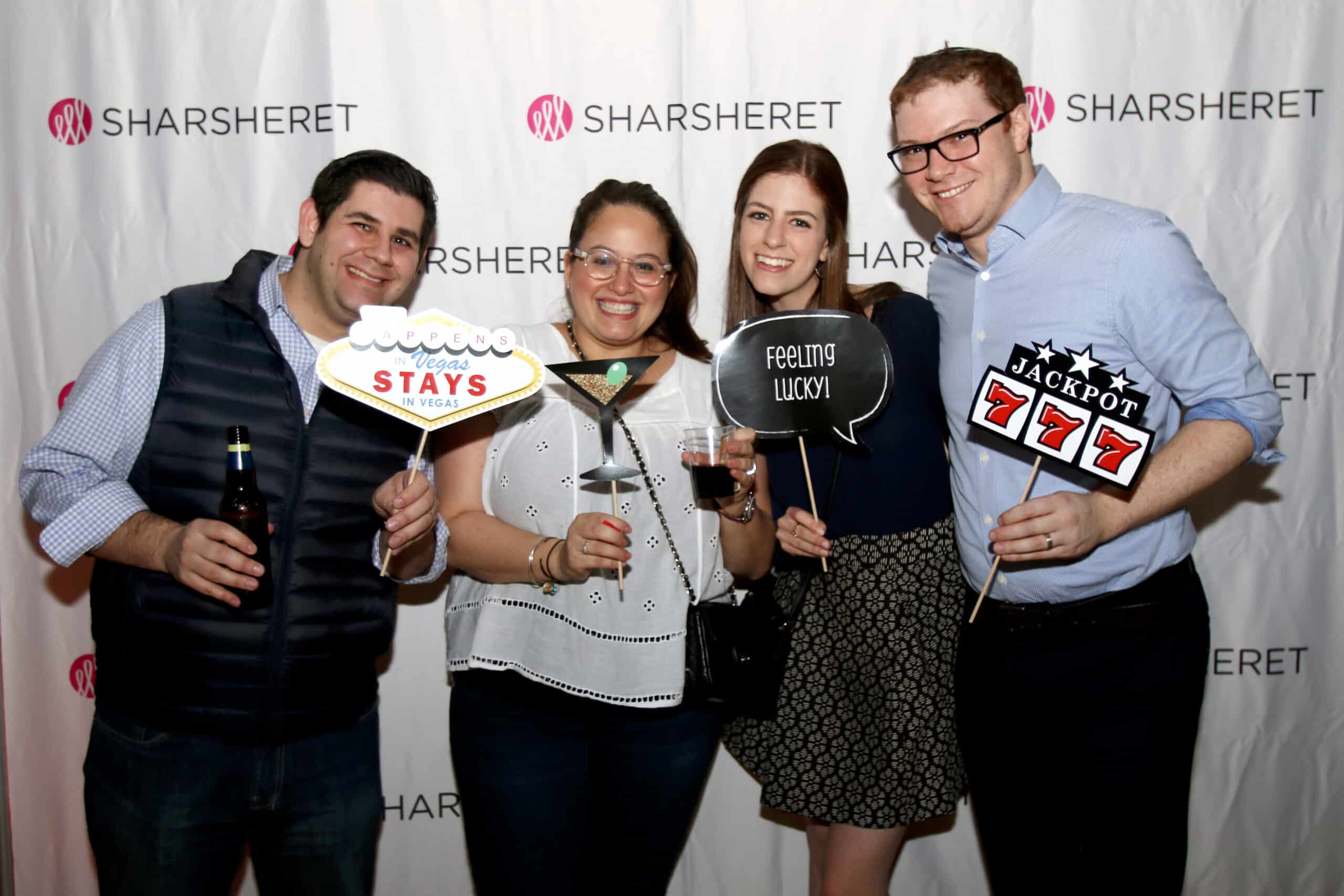 Four people stand in front of a Sharsheret-branded backdrop, smiling and holding props with messages like Feeling Lucky and Jackpot 777. One person is holding a drink and another a beer bottle.