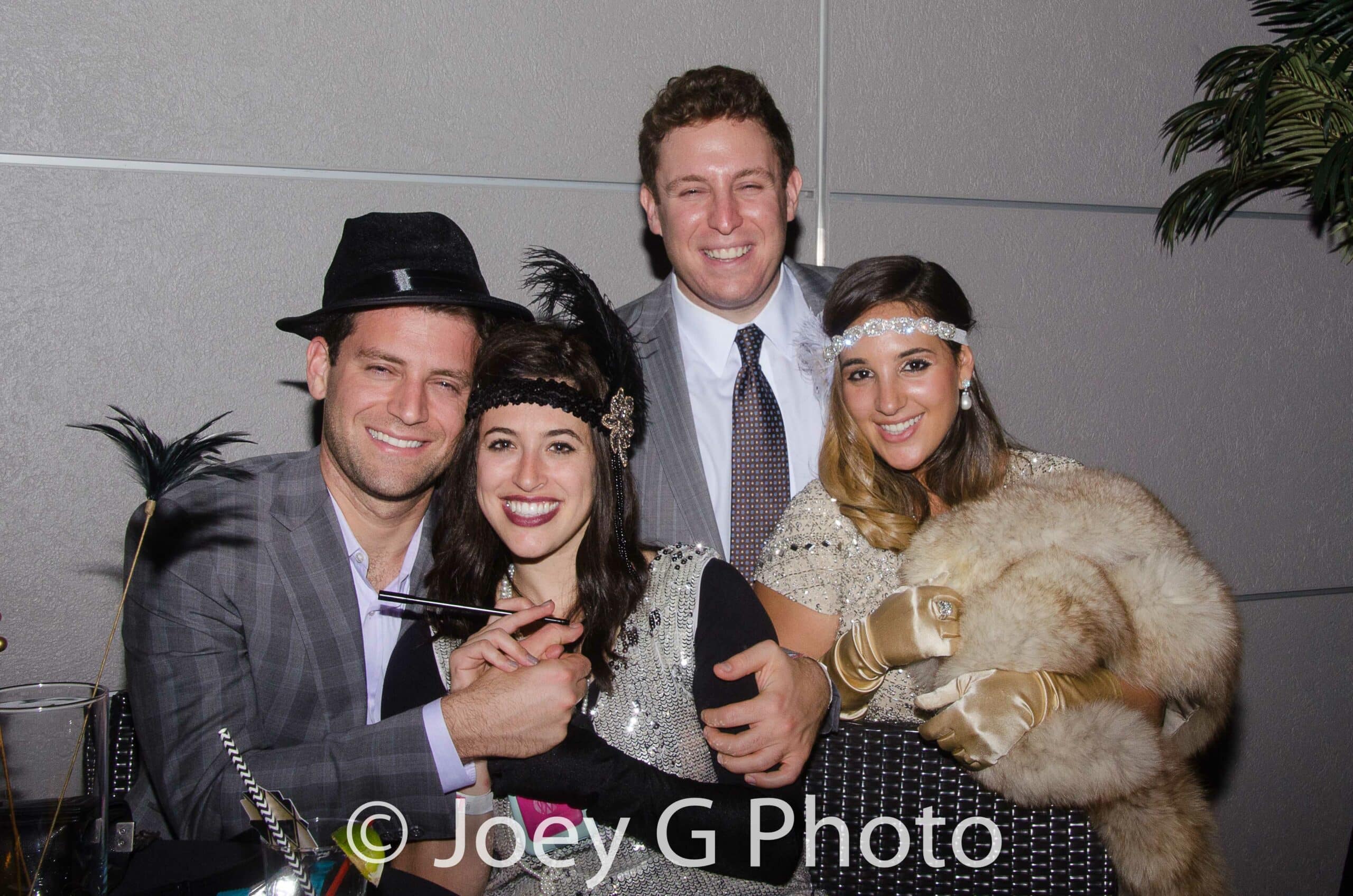 Four adults dressed in 1920s-themed attire pose together indoors, smiling at the camera. One woman wears a fur stole and gloves, another holds a cigarette holder. A photo watermark reads “© Joey G Photo.”.