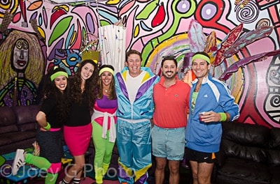 Six adults dressed in colorful, retro 1980s-style outfits pose together and smile in front of a vibrant, abstract mural with swirling patterns and whimsical figures.