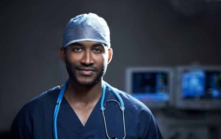 A healthcare professional in blue scrubs and a surgical cap stands indoors with a stethoscope around their neck. Medical monitors are visible in the blurred background.