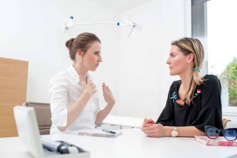 Two women sit across from each other at a desk in a bright office. One woman gestures with her hands while speaking, and the other listens attentively. A window and medical equipment are visible in the background.