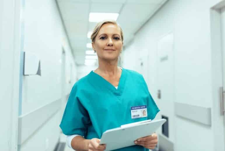 A healthcare professional in teal scrubs stands in a hospital hallway holding a clipboard, looking at the camera. A visible ID badge is attached to the uniform. The background shows a clean, well-lit corridor.