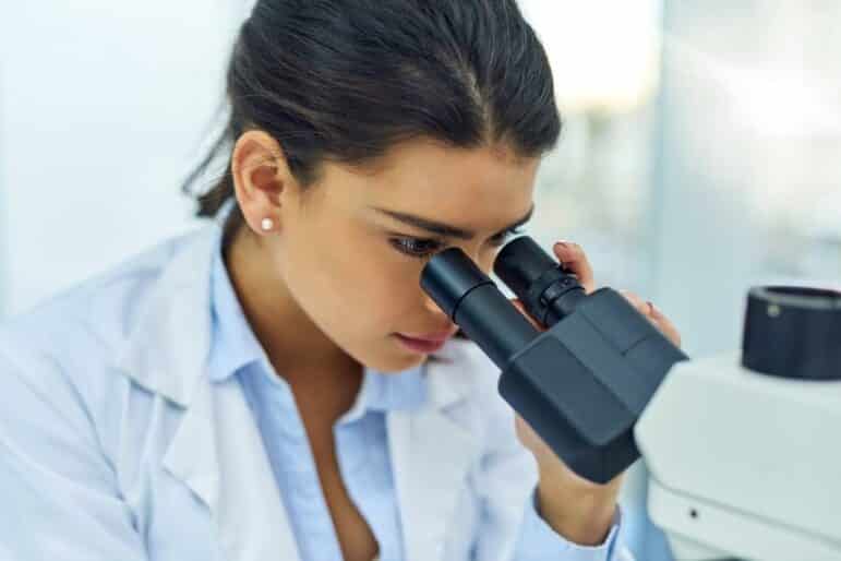 A woman in a white lab coat looks through the eyepieces of a microscope in a laboratory setting.