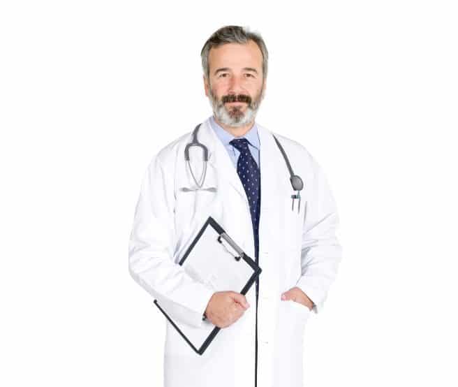 A male doctor with gray hair and a beard wears a white coat, a blue tie, and a stethoscope around his neck. He holds a clipboard and stands against a plain white background.