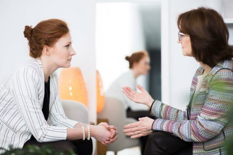 Two women sit facing each other indoors, having a serious conversation. One woman gestures with her hands while the other listens attentively, with a lamp and a plant in the background.