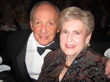 An older man in a black tuxedo and bow tie sits next to an older woman in a patterned jacket, both smiling at the camera in a dimly lit indoor setting.