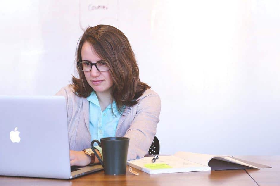 A person with long brown hair and glasses is sitting at a desk, working on a laptop. There is a black mug and an open notebook on the desk. The background is a plain whiteboard.