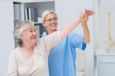 An older woman smiles while a healthcare professional in blue scrubs assists her in raising her arm during a physical therapy session in a medical office.