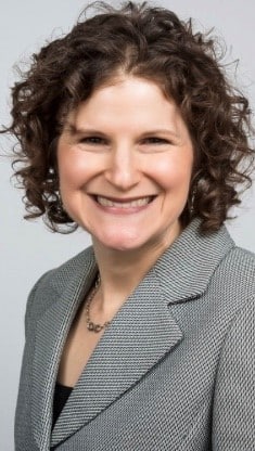 A woman with short curly brown hair wearing a gray blazer over a black top, smiling at the camera against a plain light background.