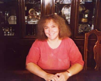 A woman with curly hair wearing a red top sits at a wooden table, smiling. Behind her is a dark wooden cabinet displaying various dishes and decorative items.