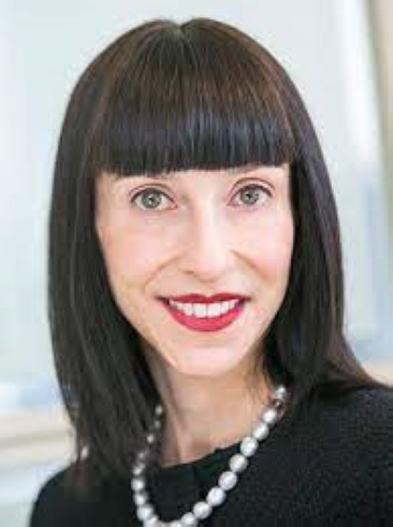 A woman with straight, dark hair and blunt bangs, wearing a black top and a pearl necklace, smiles at the camera. The background is softly blurred.
