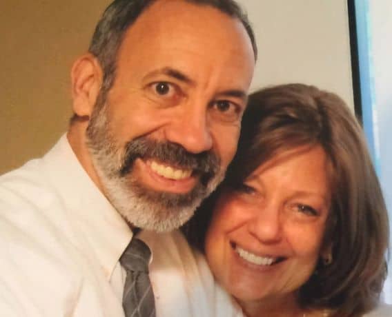 A man with short hair, a beard, and a mustache, wearing a white shirt and tie, poses closely with a smiling woman with shoulder-length brown hair. Both are looking at the camera.