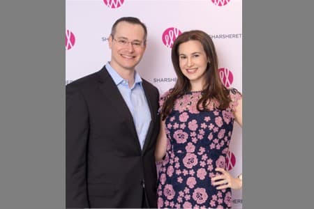 A man in a dark suit and light blue shirt stands next to a woman in a navy dress with pink floral patterns. They are smiling and posing in front of a step-and-repeat banner with pink and gray logos.