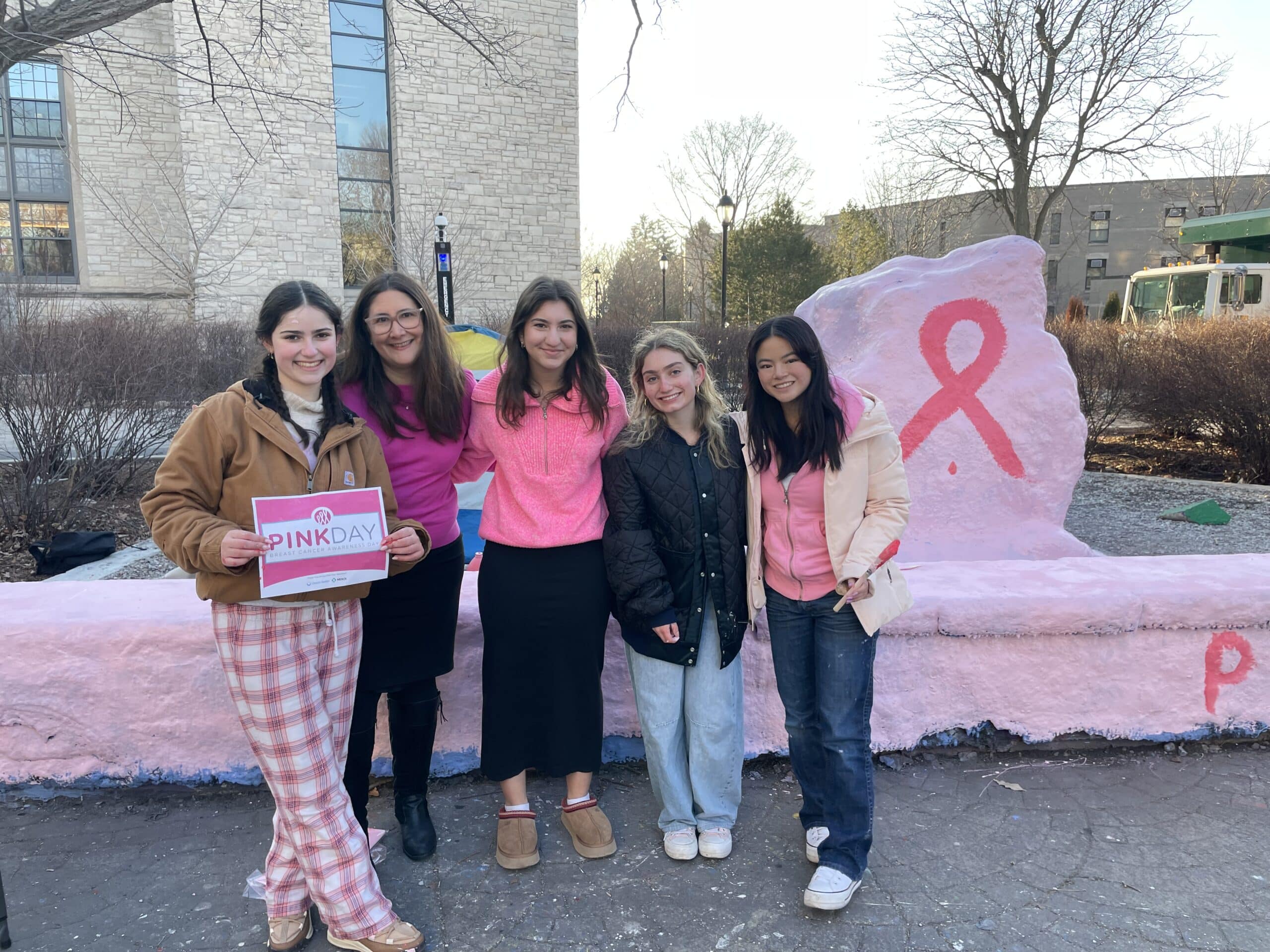 Five young women stand together outdoors in front of a pink-painted rock featuring a large pink ribbon. One holds a Pink Day sign. The group is dressed in casual clothing, some wearing pink. Trees and buildings are in the background.