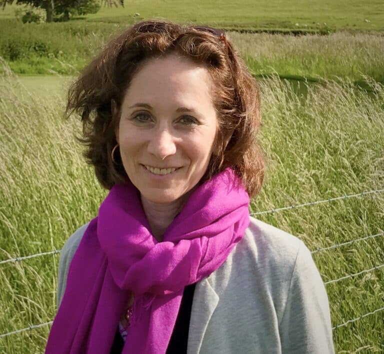 A woman with shoulder-length brown hair stands outdoors in front of tall grass and a wire fence, wearing a light gray jacket and a bright pink scarf. She is smiling at the camera.