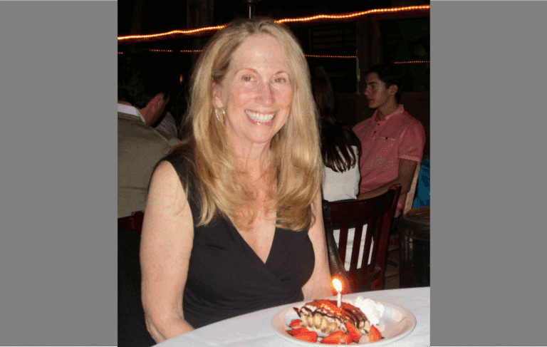 A woman with long blonde hair, wearing a black sleeveless top, sits at a table in a restaurant. In front of her is a dessert with a lit candle. Other diners are visible in the background.