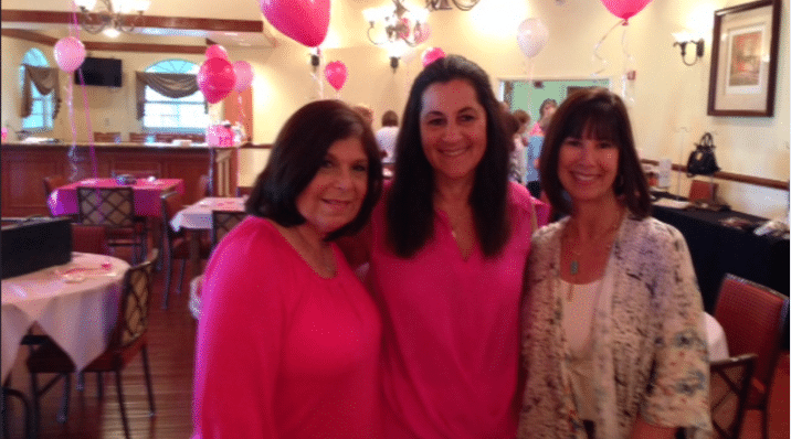 Three women stand together and smile for the camera in a decorated indoor space. Pink and white balloons are visible in the background, and the women wear pink and light-colored outfits. Tables and chairs are arranged around them.
