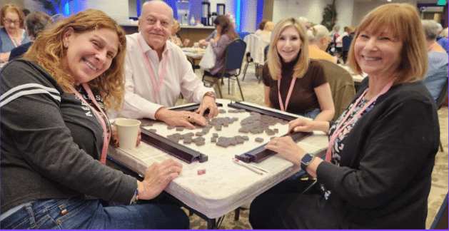 Four people sit around a table playing a tile-based game. They are smiling at the camera, wearing lanyards, and seated in a well-lit indoor setting with other people in the background.