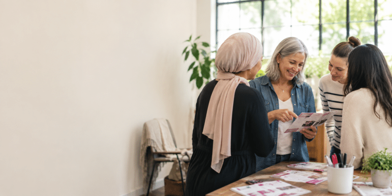 Four women stand around a table with papers, discussing community education initiatives. A plant and chair sit in the background, while large windows fill the room with natural light.