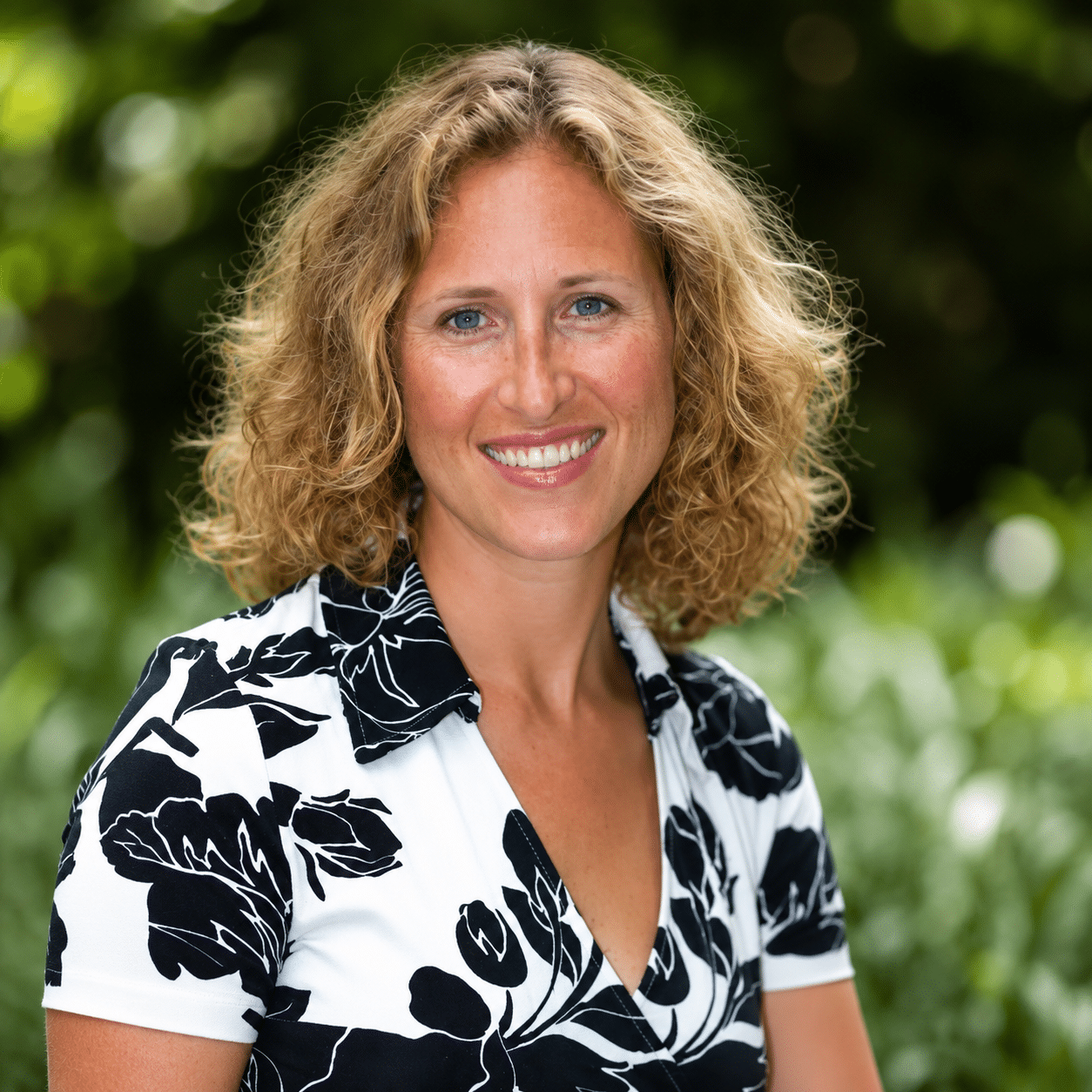 A woman with curly blonde hair is smiling outdoors. She is wearing a white blouse with a black floral pattern, and green foliage is blurred in the background.