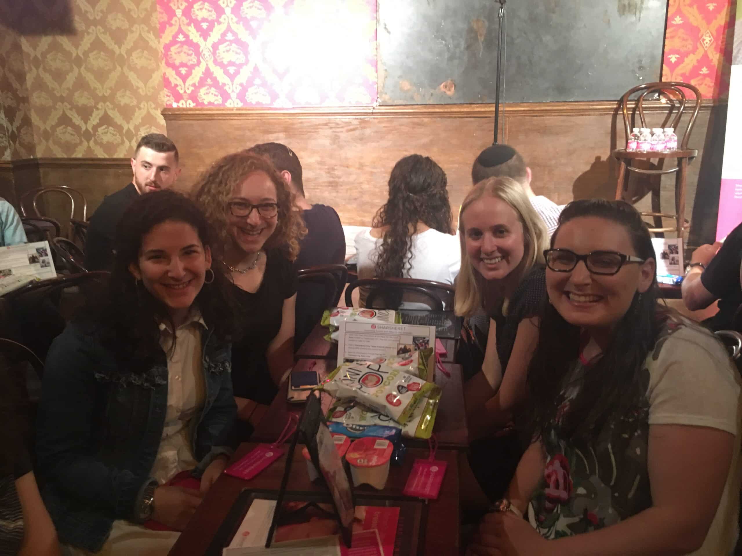 Four women sit at a table covered with brochures and various items, smiling at the camera in a dimly lit room with patterned wallpaper and a wooden chair on stage in the background.