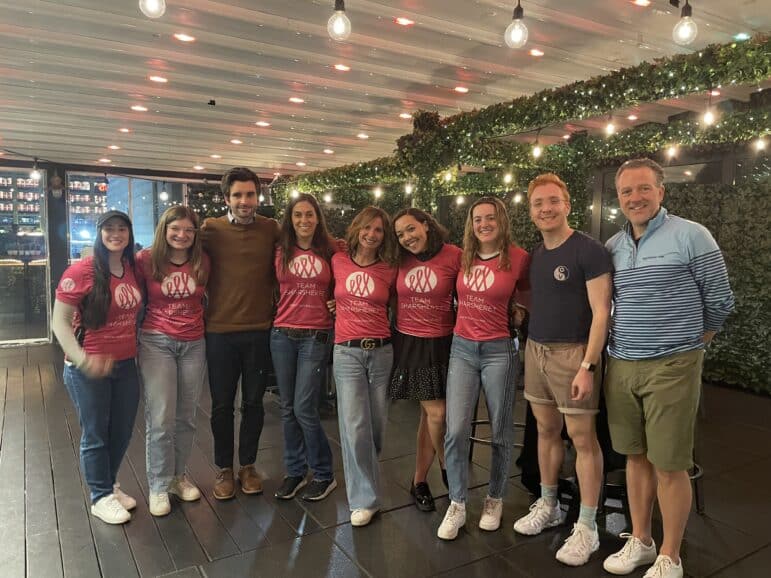 A group of nine people stands side by side on a wooden deck under string lights; six wear matching pink shirts, and three wear casual clothes. Some greenery decorates the background. All are smiling at the camera.